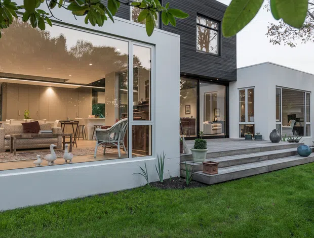 Contemporary residential rear facade opening onto a landscaped lawn and stepped terrace. The house combines smooth white rendered walls with a dark horizontal-clad upper volume, extensive floor-to-ceiling glazing, and an open-plan living area visible through the windows. Outdoor steps, potted plants, and restrained modern detailing create a strong indoor–outdoor connection.