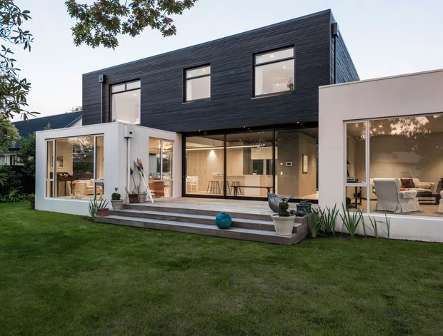 Exterior view of a contemporary two-storey residence opening onto a landscaped lawn. The home combines dark horizontal cladding on the upper level with white rendered volumes, extensive glazing, a covered outdoor terrace, and visible living and dining areas inside. Broad steps connect the interior to the garden, with restrained planting and outdoor furnishings creating a clean indoor-outdoor connection.
