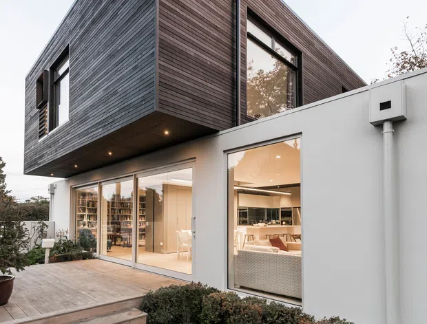 Exterior view of a contemporary two-storey home with a sharply projecting upper volume clad in dark horizontal timber boards above a white rendered lower level. Large glazed openings reveal a bright open-plan interior, while timber decking, steps, hedging and lawn form the landscaped foreground.