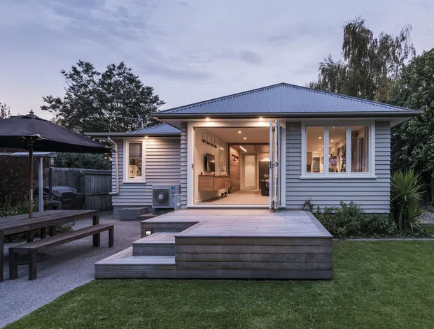 A compact single-storey contemporary home viewed from the landscaped backyard at dusk. The house has light grey horizontal weatherboard cladding, a low-pitched metal roof, white-framed windows and large glazed doors opening onto a raised timber deck with broad steps. A paved outdoor dining area with a dark umbrella and bench seating sits to the left, with lawn and established planting surrounding the extension.