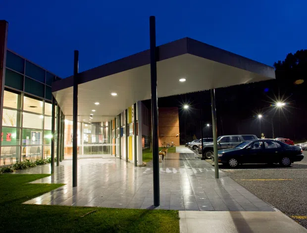 Night-time exterior view of a modern institutional or commercial building entrance. A broad flat-roofed canopy projects over a tiled pedestrian forecourt, supported by slender dark posts. Floor-to-ceiling glazing and illuminated interiors line the entrance, with brick and dark facade elements visible. A car park occupies the right side, while landscaped lawn borders the paved approach.