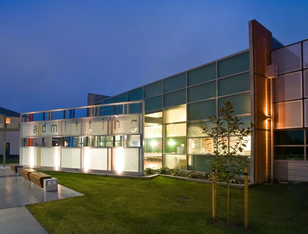 Exterior dusk photograph of a contemporary institutional or commercial building. The facade combines extensive translucent and clear glazed curtain walling, illuminated interior spaces, vertical brick-clad piers, and a projecting entrance canopy with spaced lettering. A paved forecourt, lawn, young trees, and integrated bench seating occupy the foreground.