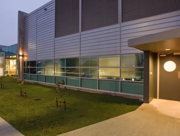 Exterior view of a contemporary low-rise commercial or institutional building at dusk. The facade combines horizontal light-coloured metal cladding, dark louvred ventilation panels, extensive gridded glazing with translucent lower panels, and a recessed entrance with a circular window. A landscaped lawn, concrete footpath and covered entry canopy frame the building.