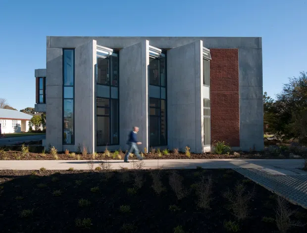 Exterior view of a compact contemporary multi-storey building with a strongly rectilinear form. Tall, narrow glazed openings are recessed between projecting grey concrete-like wall planes, with a prominent red brick feature wall on the right. A paved pedestrian path and low landscaped planting sit in front, with a neighbouring low-rise building visible to the left.