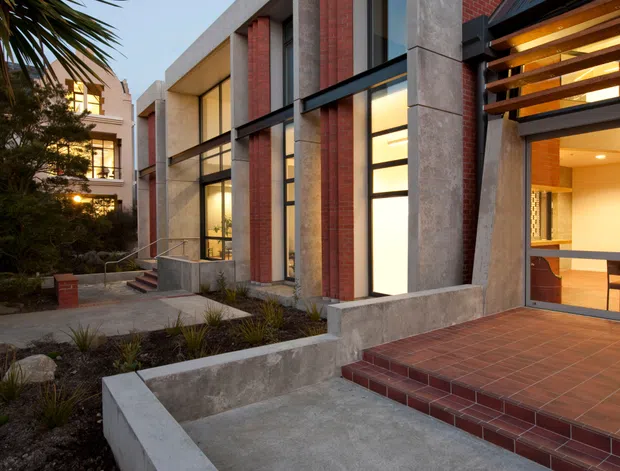 Exterior view of a contemporary multi-storey building with a rectilinear concrete frame, red brick infill walls, tall black-framed glazing, and a covered entrance. Red tiled steps and terrace paving lead through a landscaped front approach, while warm interior lighting is visible through the windows and entry.