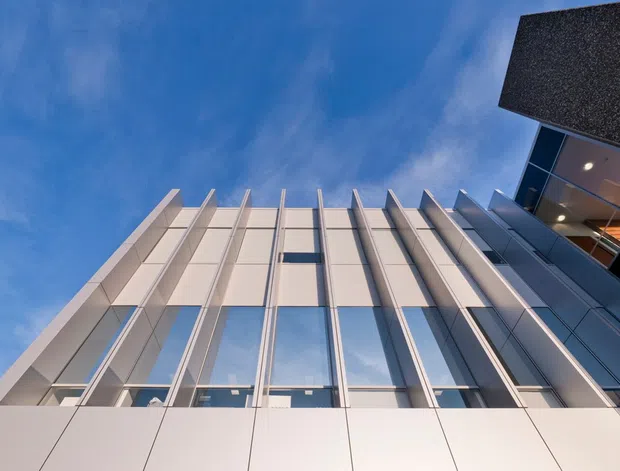 Low-angle exterior view of a contemporary multi-storey building facade, featuring white and light-grey vertical panels, extensive glazing, and projecting vertical fins. A dark overhanging roof or canopy is visible at the upper right against a blue sky.