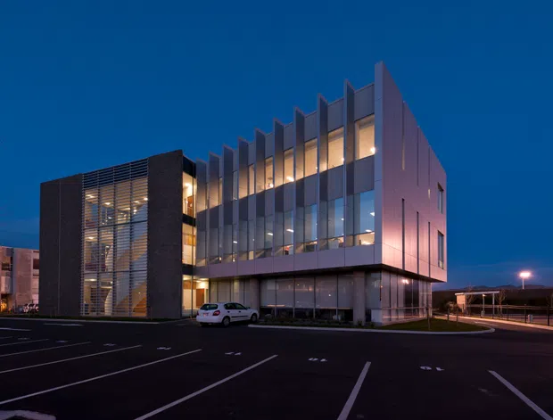 A modern multi-storey commercial building photographed at dusk, with a glazed central facade, dark brick-like end walls, projecting upper-level volumes, and a distinctive series of vertical fins along the roofline and facade. The building sits beside a marked parking area and appears warmly illuminated from within.