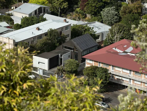 Elevated view across a leafy suburban area showing several buildings, including a prominent multi-storey structure with a broad red metal roof and a central dark pitched-roof section, surrounded by flat-roofed residential or institutional buildings and mature trees.