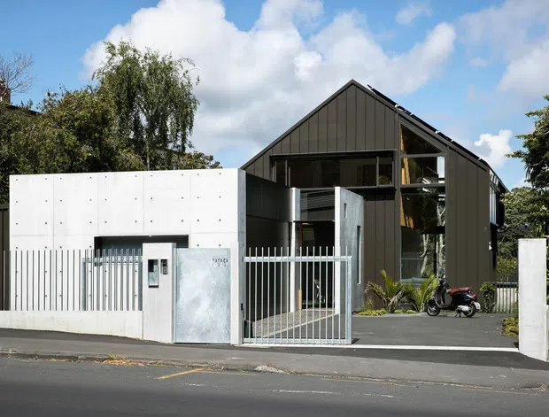 Street-facing view of a contemporary residential building with a steep gabled roof, dark vertical metal cladding, expansive glazing, and a contrasting white rendered or concrete entry volume. A metal pedestrian gate and fence enclose the paved frontage, with landscaped planting around the sides.