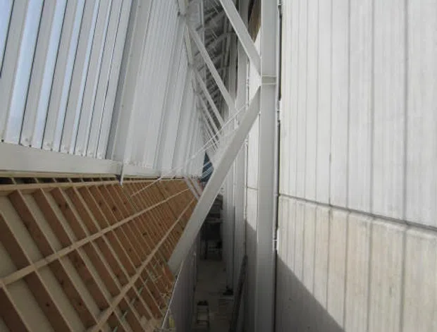 Narrow exterior service passage between large industrial building volumes, showing white ribbed metal cladding, exposed white steel bracing and roof structure, and a partially constructed timber-framed wall. The view is architectural and utilitarian, with strong linear perspective and high contrast.