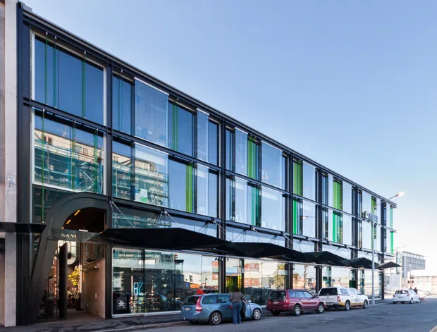 Street-level exterior view of a contemporary multi-storey commercial building with a long glazed facade, dark metal framing, and vertical green accent panels. A projecting black entrance canopy shelters a retail or office entry at ground level, with cars parked along the urban street.