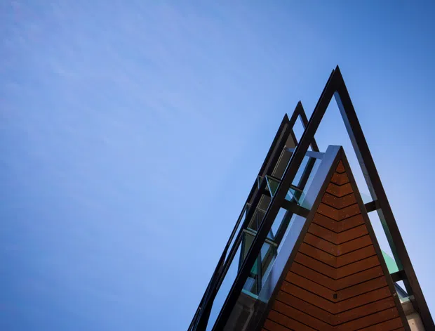 Low-angle exterior view of a sharply peaked, A-frame-like building facade against a blue sky. The structure features dark structural framing, extensive glazing, and a warm brown timber-clad triangular gable, creating a dramatic geometric composition.