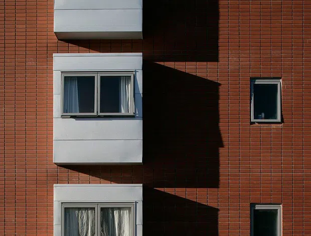 Front-on exterior view of a multi-storey brick apartment or residential building facade. Three vertically stacked white projecting window bays create strong geometric shadows across the red brick wall, with smaller dark-framed windows to the right. A metal picket fence and planted garden edge sit at the base.