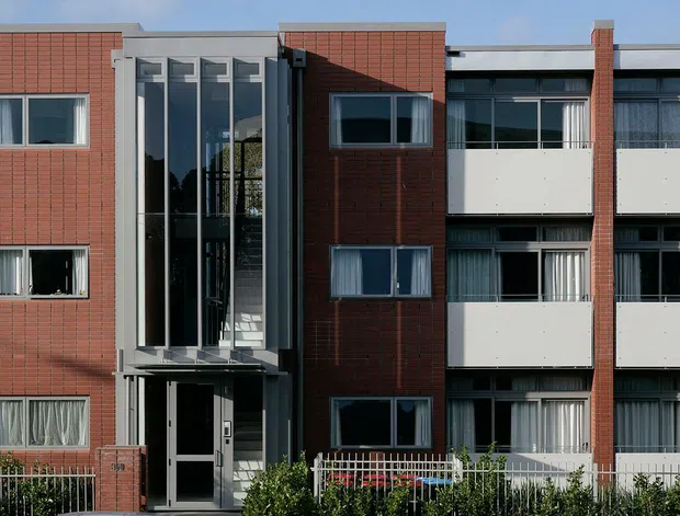 Front-facing view of a contemporary multi-storey apartment building facade. The elevation combines red brick-clad vertical sections with pale rendered or metal-panel balcony bays, dark-framed windows, and a tall glazed central stairwell or entry volume. Low planting and a metal boundary fence sit along the street frontage.