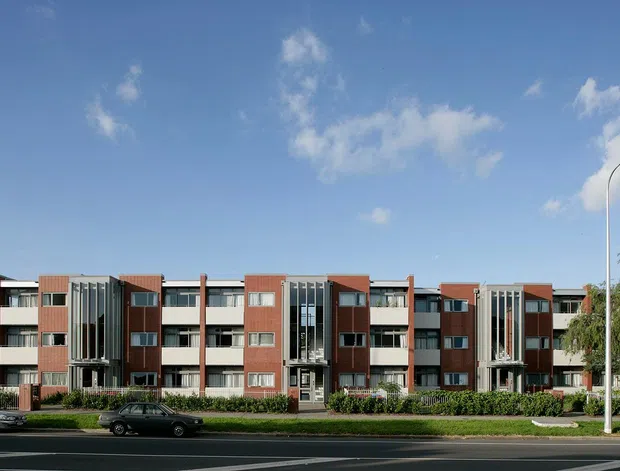 Street-level exterior view of a contemporary multi-storey apartment building with a repetitive residential facade, red-brown brick volumes, pale rendered balcony sections, extensive vertical glazing, and landscaped frontage along a broad road. The building has a mostly flat roofline and a clean, modern composition.