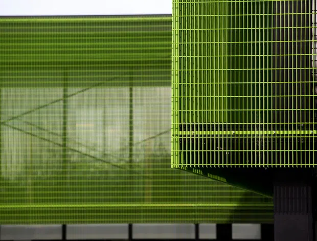 Abstract close-up architectural view dominated by layered lime-green metal mesh or louvred screening. A projecting gridded screen creates strong linear patterns and shadows over a translucent or glazed facade, with dark structural elements at the lower right. The image is highly graphic and does not clearly identify a specific room or interior.