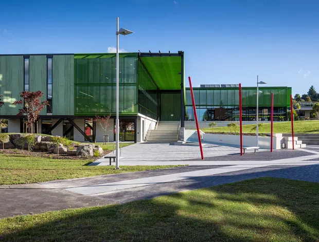 Exterior view of a contemporary multi-building campus with translucent green facade panels, extensive glazing, elevated sections, broad stairs, paved pedestrian areas, and distinctive red slender columns. The buildings sit within landscaped grounds under a clear blue sky.
