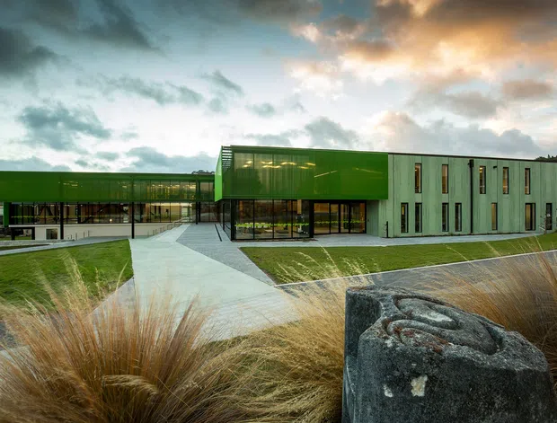 Contemporary low-rise institutional or commercial building with vivid green metal-clad volumes, extensive glass curtain walls, and a pale concrete or paved entry path. The building sits within landscaped grounds featuring ornamental grasses and a sculptural stone element, with a wooded backdrop and dramatic cloudy sky.