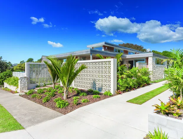 Bright exterior view of a low-slung modern residence with a flat roof, extensive glazing, pale masonry walls, and distinctive patterned breeze-block privacy screens. Tropical planting, palms, landscaped garden beds, concrete paths, and a driveway frame the home.