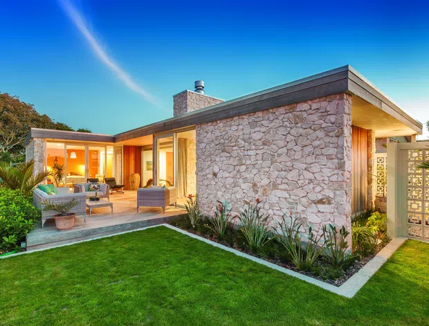 Single-storey modern mid-century-inspired home viewed from a landscaped garden at dusk. The low, flat-roofed structure combines pale irregular stone exterior walls with extensive warm-lit glazing, timber-toned interior finishes, a covered outdoor seating area, and a decorative breeze-block privacy screen. Neatly planted garden beds and lawn frame the facade.