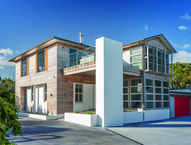 Exterior view of a contemporary two-storey residence with horizontal timber cladding, white rendered walls, broad concrete paving, a projecting upper balcony, extensive glazed openings with louvres, and an attached red garage door. The home is set among mature greenery under a bright blue sky.
