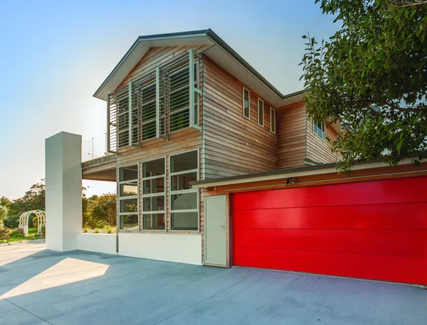 Exterior view of a contemporary two-storey residence with horizontal timber weatherboard cladding, a prominent red sectional garage door, an angled roof, and large louvred and glazed openings. The house fronts a broad concrete driveway and includes a tall white architectural wall element.