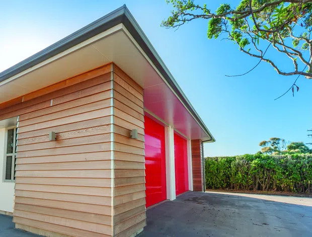 Exterior view of a contemporary single-storey building with horizontal timber cladding, a projecting flat roof, white trim, and vivid red garage or entry doors. A paved driveway and hedge-lined suburban setting are visible.