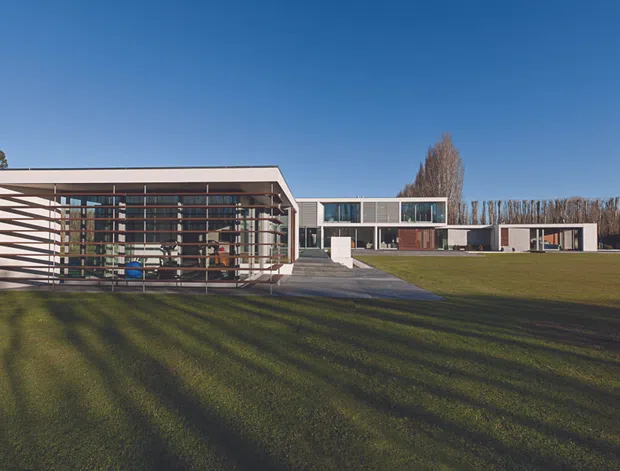 Wide exterior view of a long, low-slung modern residence set within an expansive lawn. The building has crisp white and grey rectangular volumes, extensive floor-to-ceiling glazing, and a prominent projecting horizontal timber or metal brise-soleil screen across the left pavilion. A paved path connects the lawn to the entry, while tall rows of trees form the background. Clear blue sky and strong low-angle shadows emphasize the horizontal composition.