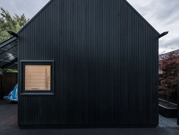 Front exterior elevation of a compact, simple gable-roofed building clad entirely in dark vertical boards. A small square window with timber venetian blinds sits low on the facade, while a covered side structure and landscaped boundary are partially visible. The image has a moody, minimalist architectural character under an overcast sky.