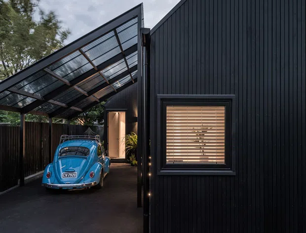 Exterior view of a dark, contemporary residential building with steeply angled roof geometry and vertical black cladding. A transparent slatted-roof carport shelters a vintage blue car, while a small window with timber venetian blinds and a lit entry passage are visible. The scene is photographed at dusk and has a dramatic, minimalist character.