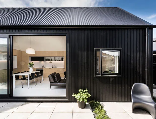 A contemporary single-storey detached building with a steeply pitched corrugated metal roof and black vertically ribbed cladding. Large sliding glass doors reveal a bright open-plan kitchen and dining area, with a small living seating area inside. The exterior has a pale tiled terrace, a narrow planted strip, and a sculptural dark outdoor chair. A neighbouring roofline is visible at the side.