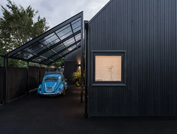 Exterior view of a compact contemporary building clad in dark vertical ribbed panels, with a large angled translucent-roof carport and a vintage blue car parked beneath it. A small window with timber venetian blinds punctuates the facade, while dark fencing and mature planting frame the setting.