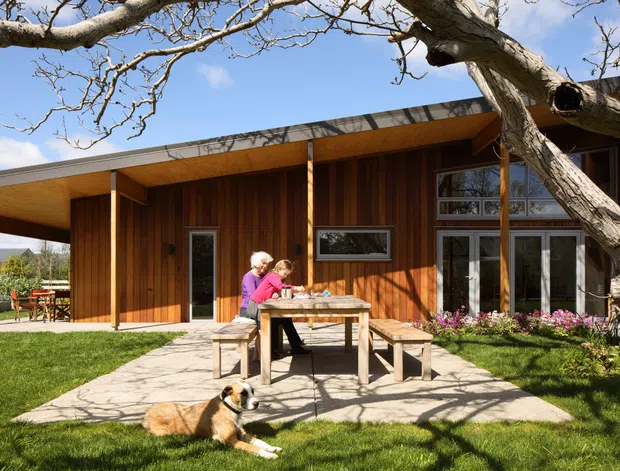 A single-storey modern home with a broad low-pitched roof and extensive warm timber cladding, photographed from a landscaped garden. A covered outdoor dining patio with a timber picnic table sits in front of large glazed doors and windows. Mature tree branches frame the scene, with lawn, planting, and people and a dog in the foreground.