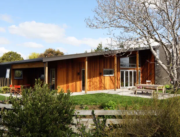 Single-storey modern timber-clad house set within a landscaped garden. The long, low building has a shallow flat or low-pitched roof, dark-framed windows, extensive warm vertical timber wall cladding, and a covered outdoor terrace with simple bench seating. A timber boardwalk and native-style planting occupy the foreground.