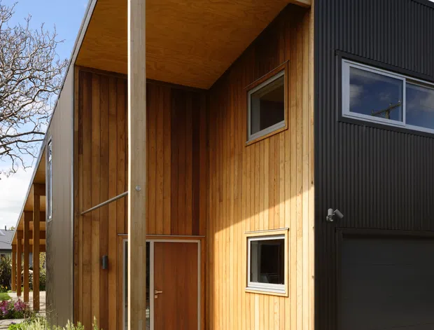 A contemporary multi-storey residential facade viewed from outside, featuring a steep gabled roof form, black vertically ribbed exterior cladding, and a recessed timber-clad entry porch. The porch has a large timber soffit, exposed structural post, timber wall lining, glazed entry door, and small horizontal windows. A dark garage door occupies the right side, while landscaped planting and a textured driveway sit in the foreground under a bright sky.