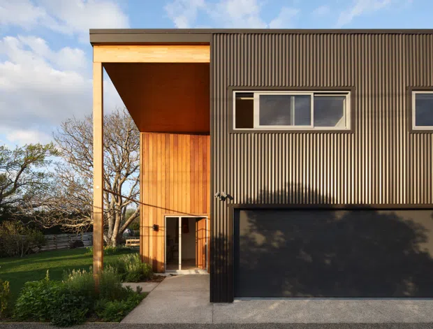 Exterior view of a contemporary two-storey house with a strongly geometric facade. The building combines dark vertically ribbed cladding with warm timber lining at a tall recessed entry and upper-level overhang. A wide dark garage door occupies the lower right, while narrow horizontal windows and a timber-framed entry provide contrast. Landscaping, lawn, mature trees, and a concrete path surround the frontage.