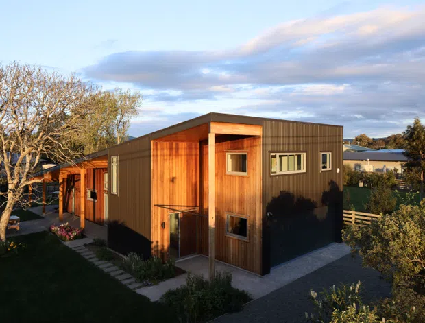 Exterior view of a compact contemporary two-storey house with a rectilinear flat-roofed form. The facade combines warm vertical timber cladding with dark corrugated or ribbed metal panels, large timber-framed openings, and a small paved entry path. The home sits in a landscaped suburban setting with mature trees and neighbouring buildings visible around it.
