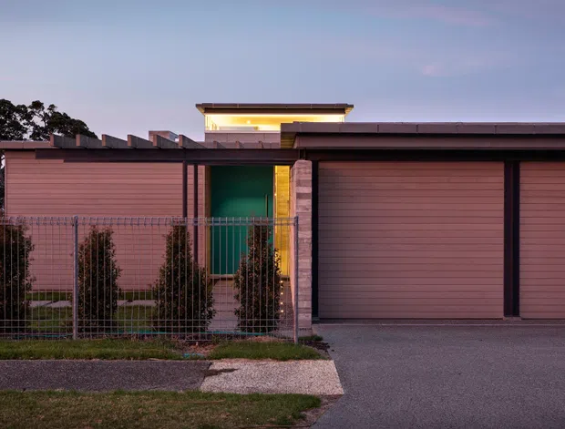 Street-level dusk view of a low, contemporary residential facade with horizontal muted-terracotta cladding, two wide garage doors, a recessed green entry door, brick piers, warm linear lighting, and a simple flat roofline. A metal front fence, narrow planting, driveway, and pavement are visible in the foreground.