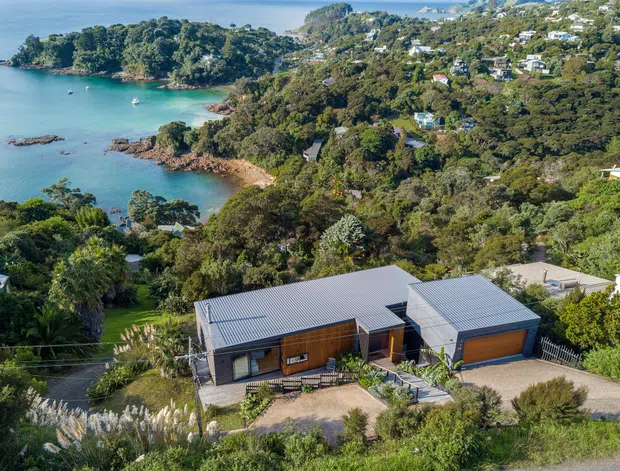Aerial view of a contemporary single-storey residence set into a heavily vegetated coastal hillside. The home has low-pitched grey metal roofs, dark timber-clad walls, extensive glazing, and an attached garage with a broad timber-toned door. A landscaped driveway and garden surround the house, with a sheltered turquoise bay, rocky shoreline, and additional hillside homes visible beyond.