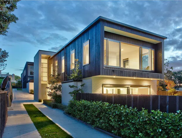 Contemporary multi-level residential house viewed from the street at dusk, featuring dark vertical cladding, projecting box-like forms, large illuminated windows, pale masonry at the lower level, and a landscaped front boundary with hedging and a pedestrian path.