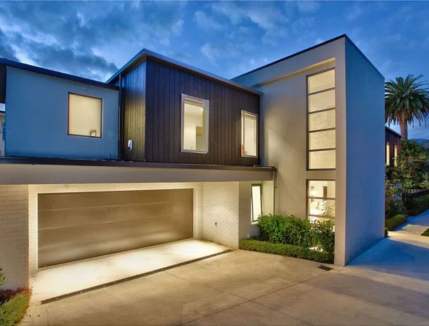Twilight exterior view of a contemporary multi-level residence with a flat and low-pitched roof composition, white rendered walls, dark vertical timber-look cladding, and extensive glazed openings. The house includes an integrated double garage, tall stairwell glazing, landscaped planting, and a concrete driveway with a pedestrian path.