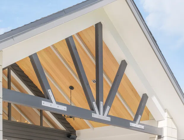Close-up exterior architectural view of a contemporary gabled roof or canopy, featuring exposed dark structural steel beams and brackets beneath a warm timber-lined soffit. White fascia and wall cladding frame the structure, with blue sky visible beyond.
