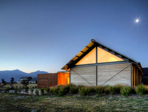 Exterior view of a contemporary gable-roofed building at dusk, with a timber-clad facade, large triangular glazed gable glowing with warm interior light, dark structural framing, and a landscaped grass foreground. Mountain silhouettes and neighbouring buildings are visible in the distance.