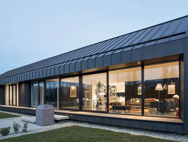 A long, single-storey contemporary building photographed at dusk, with a steep pitched metal roof and dark charcoal exterior cladding. A full-height glazed facade reveals a warmly lit open-plan living area with lounge seating, shelving, and pendant lighting. The building sits within a landscaped gravel-edged garden, with a strong linear roof and facade composition.