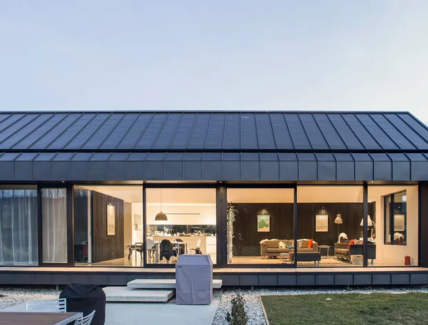 Exterior view of a low, elongated contemporary house with a dark standing-seam metal gabled roof and dark vertical-clad facade. Full-height glazed sliding doors reveal a warmly lit open-plan kitchen and living area, opening onto a simple paved terrace and lawn.