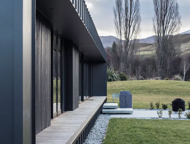 Exterior architectural photograph of a long, linear contemporary building with dark cladding, extensive vertical glazing, and a projecting upper volume forming a covered timber walkway or deck. The building sits beside a carefully landscaped lawn with gravel drainage strips, a pale paved path, outdoor furniture, tall slender trees, and mountainous rural terrain in the background. The composition emphasises strong horizontal and vertical lines and a restrained dark neutral palette.