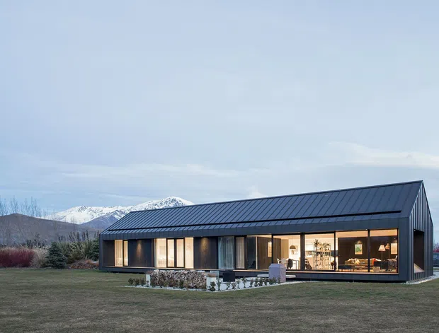 A single-storey contemporary home set in an open rural landscape, photographed at dusk. The building has a long rectangular form with a steep gable roof clad in dark standing-seam metal, extensive warm-lit glazing along the front facade, dark exterior cladding, and a small covered entry or terrace. Stacked firewood and sparse landscaping sit around the house, with mountains visible in the background.