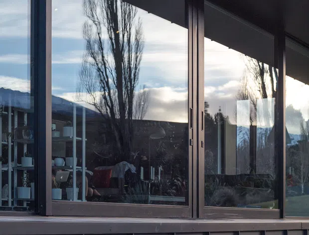 Exterior architectural photograph of a contemporary dark-clad building with extensive floor-to-ceiling glazing, metal-framed windows and doors, and projecting concrete entry steps. The glazing reflects tall trees, sky, and distant mountains while revealing a furnished interior beyond. The composition emphasises the facade, glazing, and layered entrance platform.