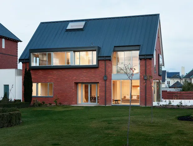Exterior view of a contemporary two-storey brick house with a steep dark standing-seam metal gable roof, roof skylight, large projecting and floor-to-ceiling windows, and a landscaped front lawn. The red brick facade is paired with dark-framed glazing and a simple modern form.