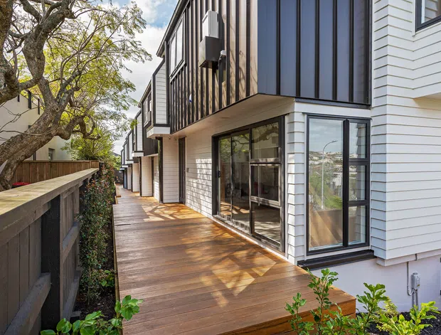 Exterior view of a contemporary multi-level residential building, showing a timber deck and narrow side walkway beside dark fencing and landscaping. The facade combines white horizontal weatherboards with dark vertical cladding, large black-framed glazing, projecting balconies, and a covered outdoor living area.